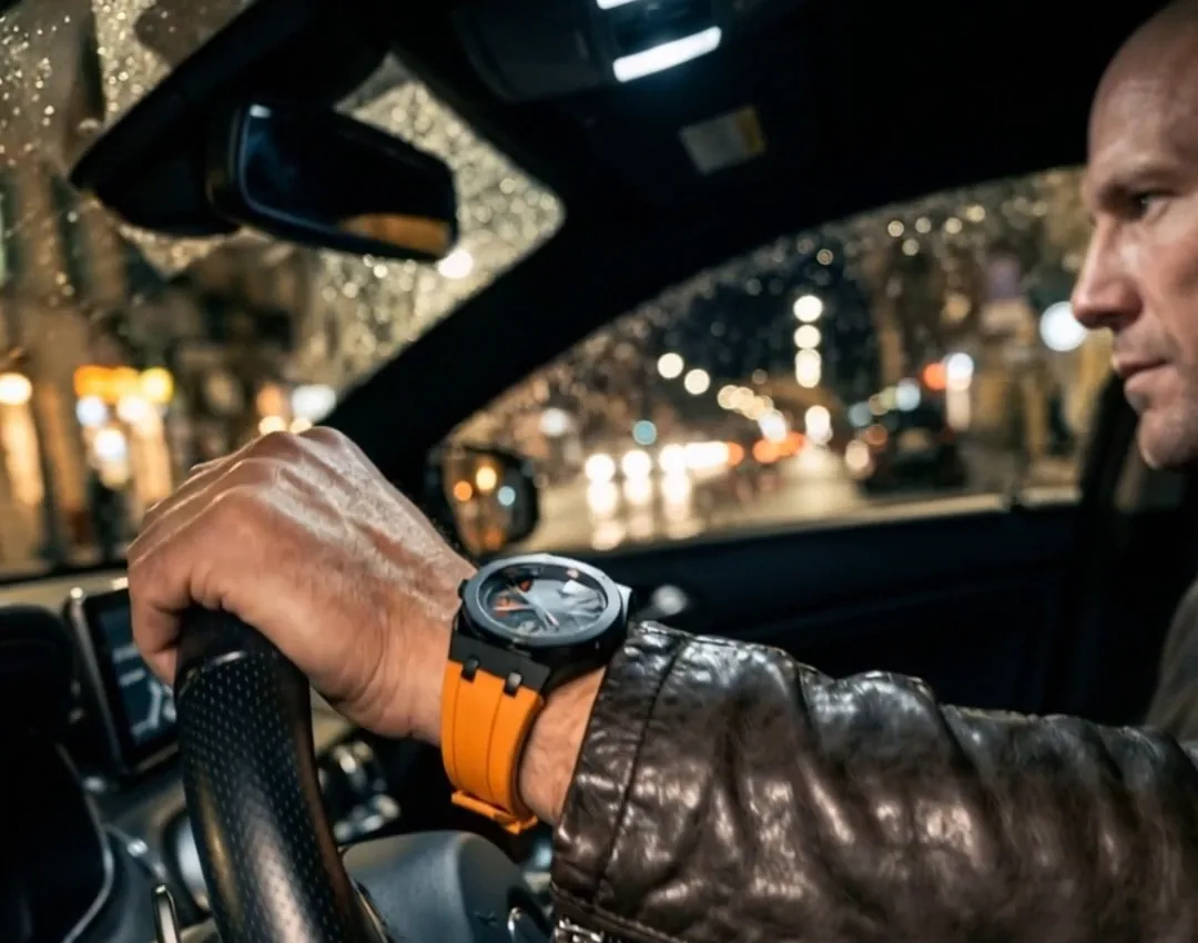 Close up inside a car at night in the rain. The bald man is in profile at the right edge of the frame, looking down the street. One hand grips the steering wheel with a black and orange skeleton watch on the wrist, and city lights glow as blur behind the wet window. AI generated.